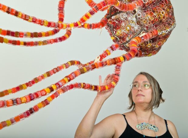Artist Kait Rhoads stands below one of their pieces; a colorful glass jellyfish